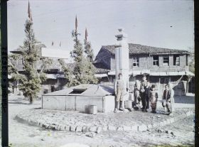 Image représentant Enfants et homme à une fontaine ; la rue est décorée d'arbres ou de mâts habillés de feuillages et coiffés de drapeaux turcs ; entre 2 mâts, une inscription sur un panneau