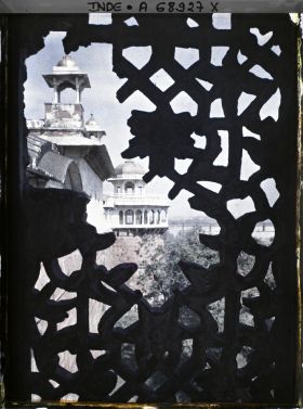 Image représentant Vue sur l'extérieur de l'enceinte du fort Rouge (Lal Qila) depuis une fenêtre ornée de jalis ; on aperçoit la toiture du palais privé (Khas Mahal) surmontée d'un chhatri et au fond, la tour du Jasmin (Musamman Burj)