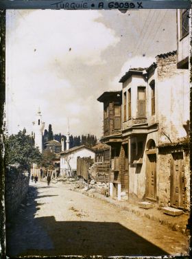 Image représentant Une rue bordée de maisons en dur et en bois,  deux petits minarets à l'arrière plan