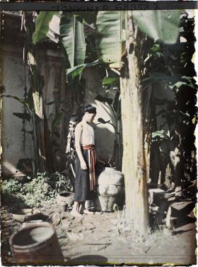 Image représentant Une jeune fille en cache-sein et turban dans un coin de cour planté de bananiers