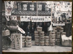 Image représentant Epiciers à l'entrée de leur boutique, devant laquelle sont exposés des fûts de bière, de "miso" (pâte de haricots de soja fermentée), de saké et du charbon de bois