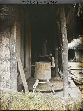 Image représentant Une jeune fille actionnant un moulin à décortiquer le paddy, dans une cour de ferme après la moisson du riz