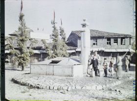 Image représentant Enfants à une fontaine ; la rue est décorée d'arbres ou de mâts habillés de feuillages et coiffés de drapeaux turcs ; entre 2 mâts, une inscription sur un panneau