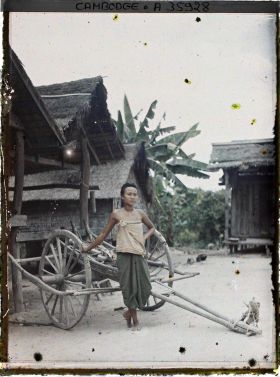 Image représentant Une femme d'ethnie badak, en sampot large, appuyée à une charrette devant un groupe d'habiations