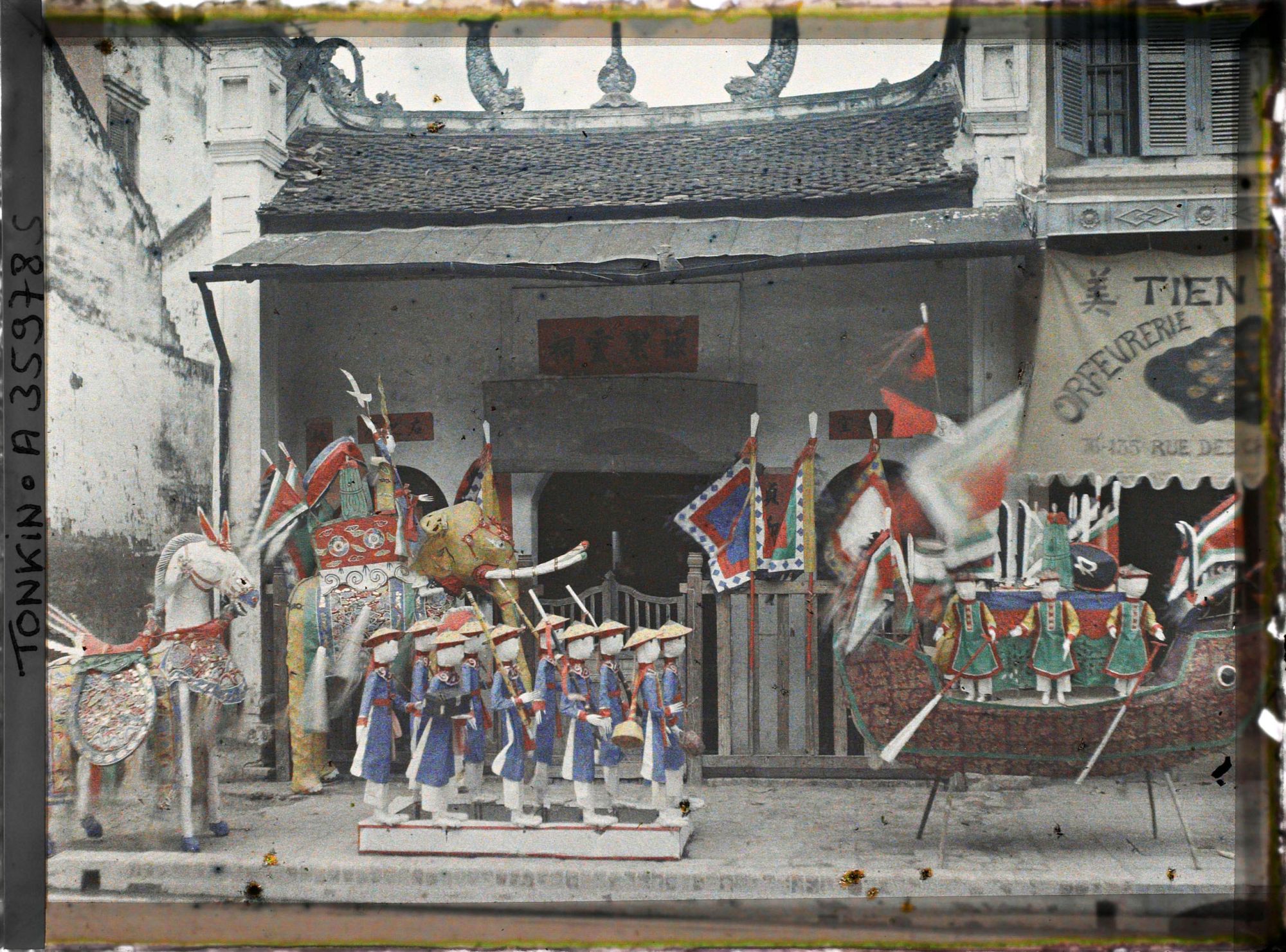 Image représentant Une exposition d'objets votifs en papier (personnages, embarcation, cheval, éléphant) devant un petit temple