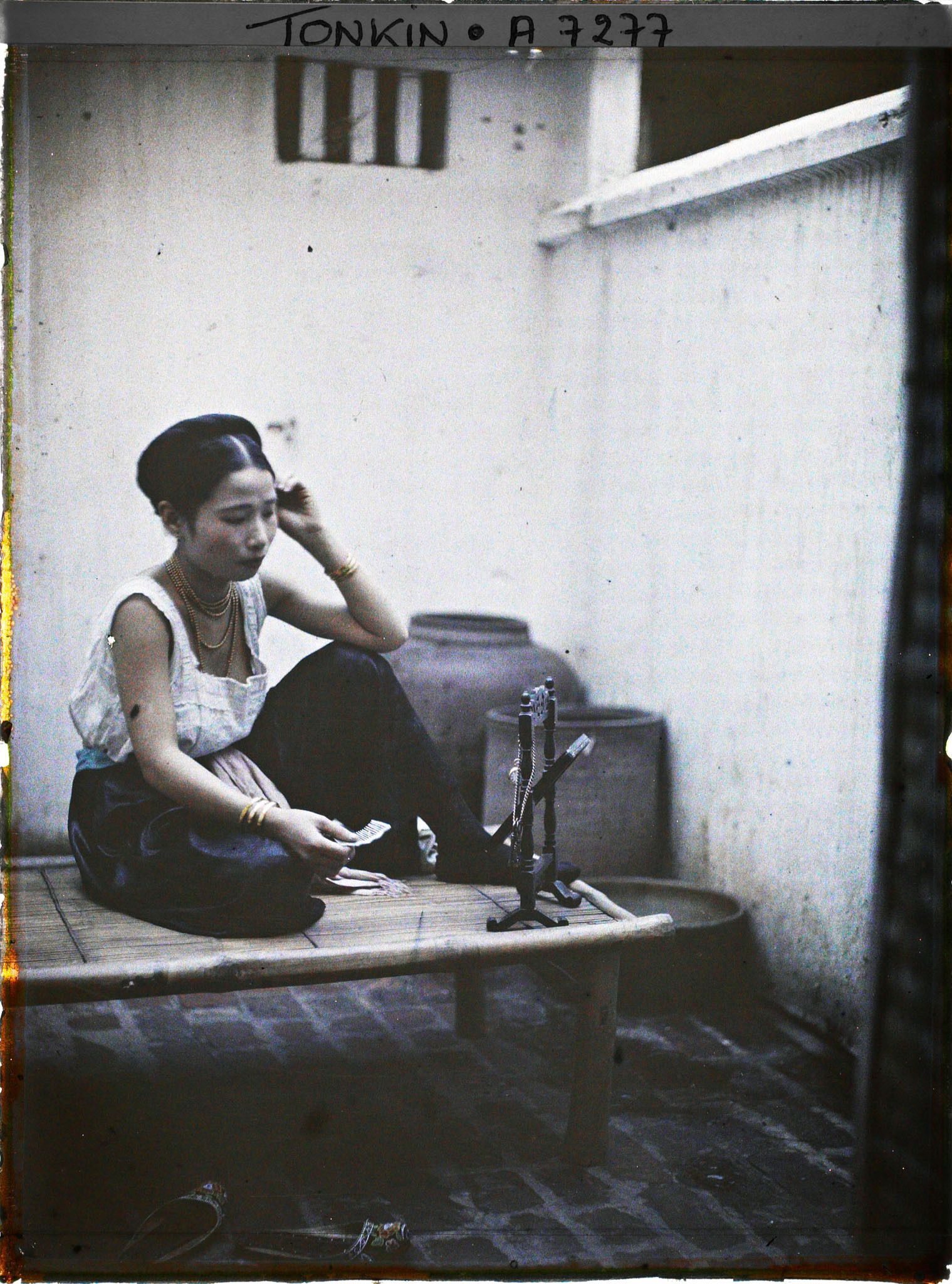 Image représentant Une jeune élégante de classe aisée à sa toilette, assise sur une banquette en bambou dans la cour intérieure de son habitation