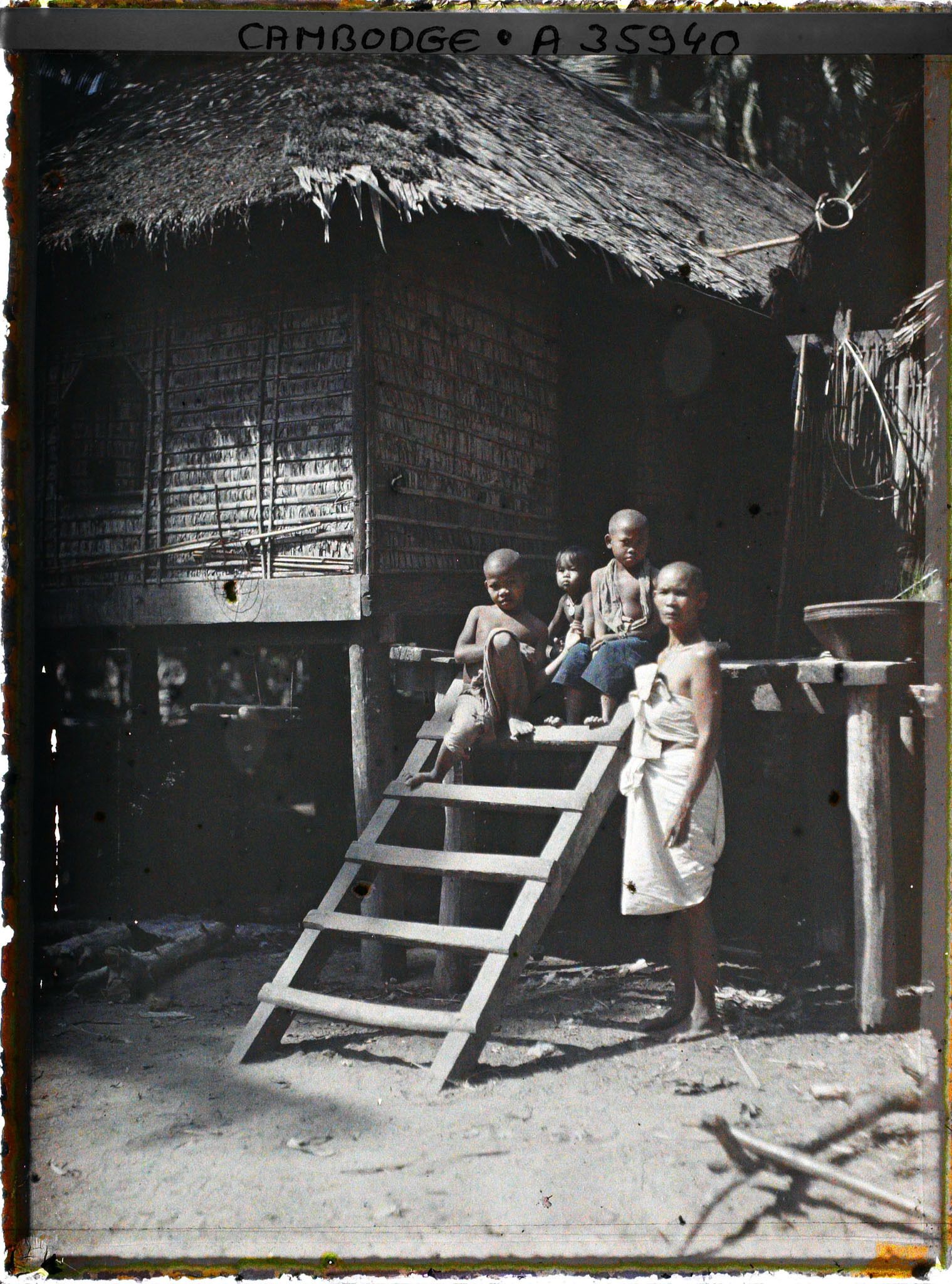 Image représentant Une femme en deuil et des enfants près de l'échelle permettant d'accèder à une maison sur pilotis